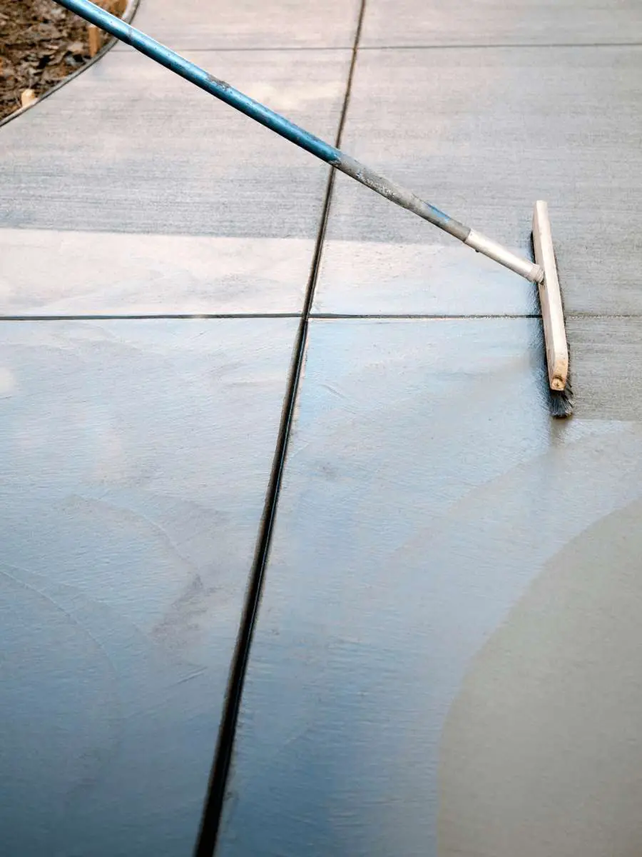 A person is smoothing freshly poured concrete on a sidewalk in Coral Gables, FL, using a long-handled trowel. The concrete, potentially from Dade Decorative Concrete, is still wet and has visible tool marks, indicating it is being worked on for that perfect decorative finish.